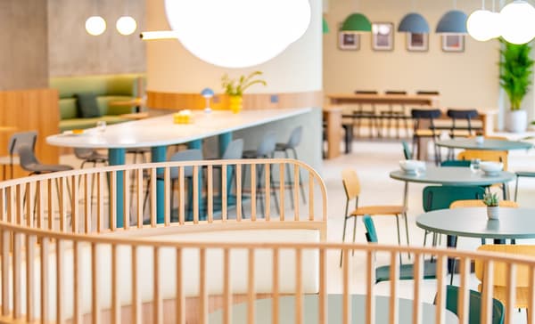 Wide view of a bright and spacious communal shared space. The space is furnished with a mix of long white bar-height tables, small round tables, and semi-circular booths with light wood slat railings.