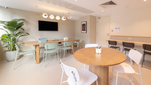 Multi-purpose room with a long wooden table for meetings or group work, a smaller round table, mint-green and white chairs, and a wall-mounted monitor.