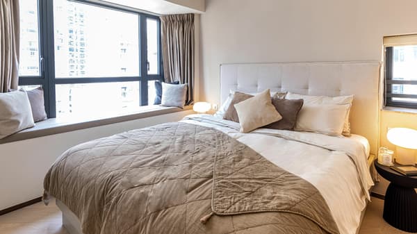 Bright master bedroom featuring a queen bed with a tufted headboard, a cushioned window seat, and a large window with city views.