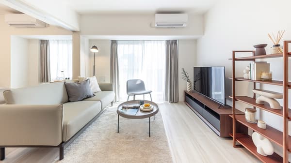 Bright modern living room with a light gray sofa, dark wood entertainment unit, and a decorative open shelving unit, facing a large window and balcony.