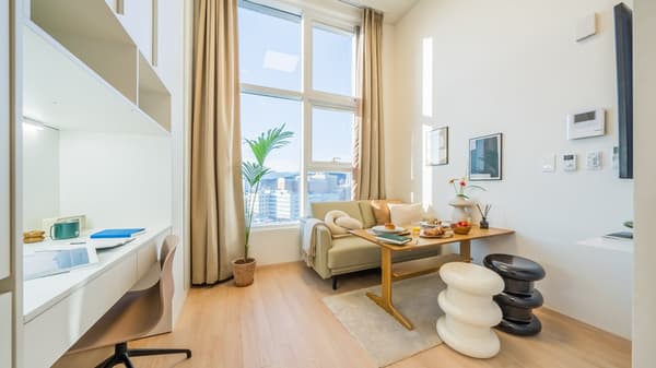 View of an apartment living area featuring a sofa, dining table, two spiral stools, and a bright built-in desk area, under tall windows with a city view.