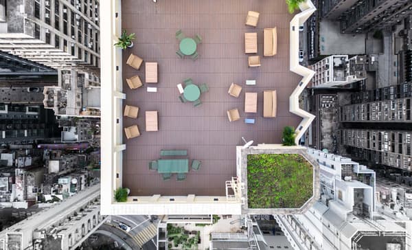 Aerial view of a rectangular, wood-decked rooftop terrace with various outdoor seating arrangements and green patches, surrounded by tall city buildings.