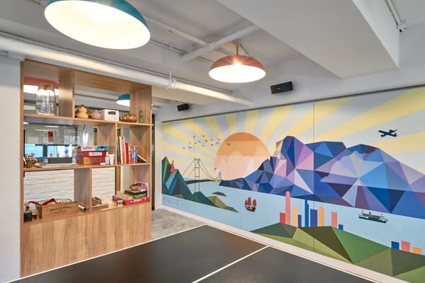 Common area recreation room featuring a ping pong table in the foreground, a wooden bookshelf divider, and a large, colorful geometric wall mural depicting a stylized Hong Kong skyline at sunrise.