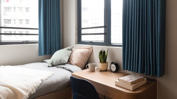 A compact single studio bedroom featuring a bed with pastel pillows, a wooden desk with a laptop, a plant and an alarm clock, and two large windows with navy blue curtains.