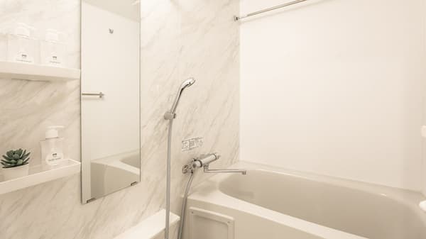 Clean, white bathtub and shower area in a Japanese unit bathroom, with white marble-effect wall panels and a mirrored wall shelf.