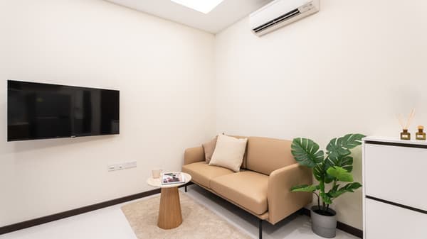 Compact living area featuring a tan two-seater sofa, a small round wooden side table, a wall-mounted television, and a potted plant.