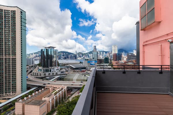 Panoramic view from an outdoor balcony, showing a modern Hong Kong cityscape, including a major highway interchange, several tall residential towers, and the distant skyline against a bright blue, cloudy sky.