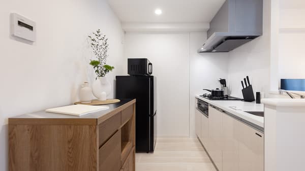 Narrow, minimalist galley kitchen featuring white cabinets, a black fridge, microwave, gas cooktop, and a light wood sideboard with decor.