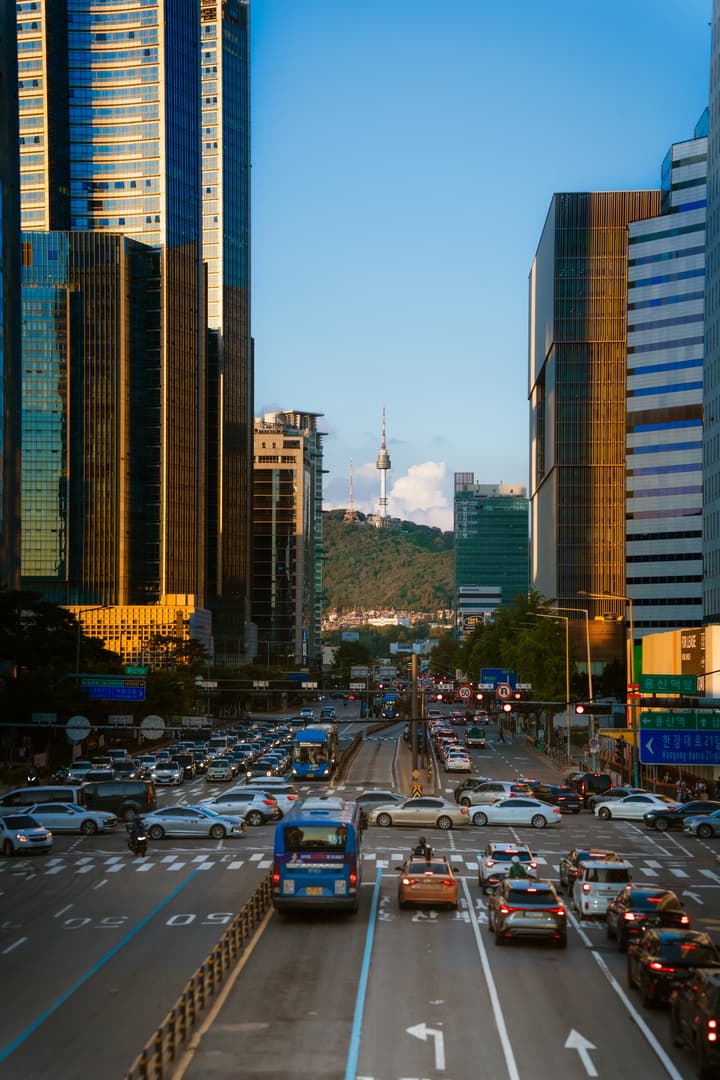 A bustling street scene in Seoul, South Korea, with high-rise modern buildings framing the street and the N Seoul Tower visible on a distant green hill.