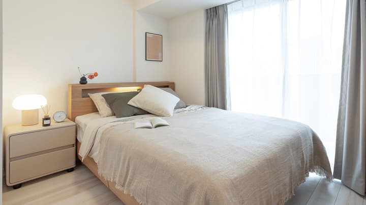 Bright primary bedroom featuring a double bed with a light wood frame, a beige nightstand with a lamp, and a large window with sheer and gray blackout curtains.