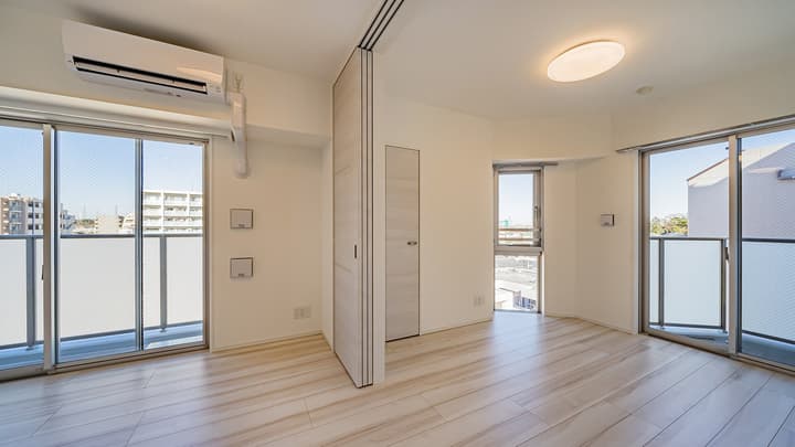 Bright living room with light wood-style flooring, a wall-mounted air conditioner, a small window, two sliding glass doors leading to separate balconies, and a central light wood room divider panel.