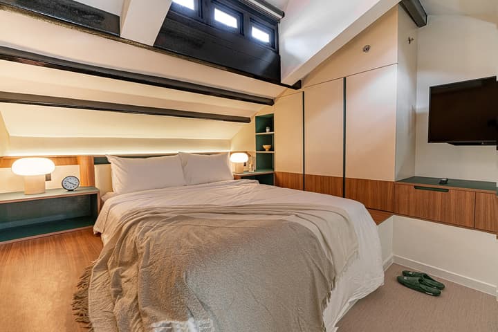 Upper level of a loft suite featuring a cozy queen bed, bespoke teal and wood cabinetry, and an angled, vaulted ceiling with dark beams.