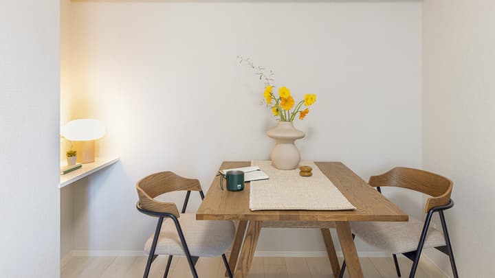 Close-up of the apartment's dining area featuring a wooden table with a vase of yellow flowers, two modern wooden chairs, and a small built-in shelf with a desk lamp in Monzennakacho.