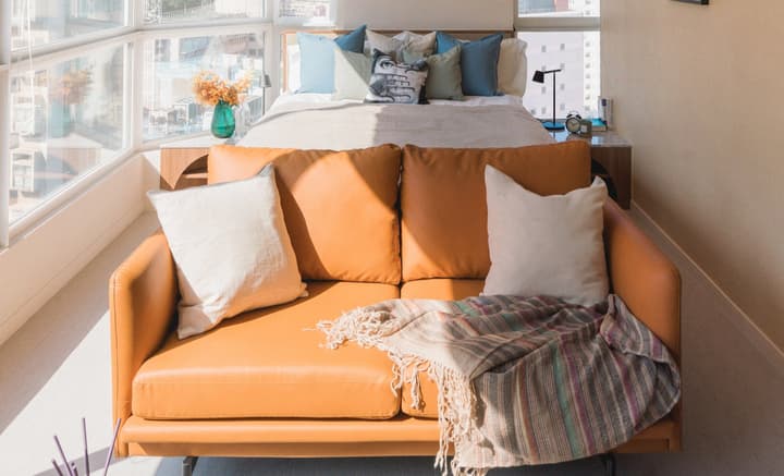 Sunlit studio apartment featuring a tan leather sofa, cozy bed with colorful cushions, and large windows offering city views in Hong Kong.