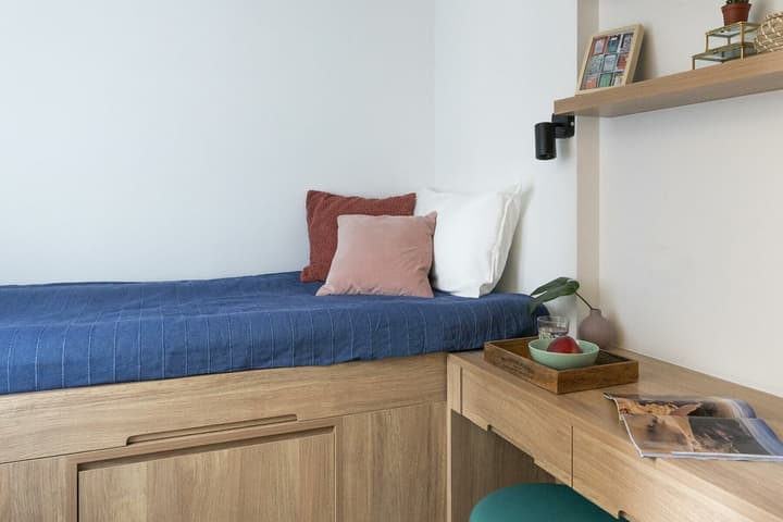 Close-up of a compact single bed in a studio, featuring a blue quilted bedspread, various pillows, a built-in wooden frame with storage drawers, and a connected wooden desk with a small tray holding fruit and a drink.