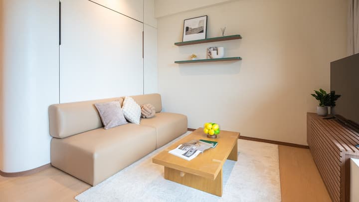 Murphy bed concealed as a white wall panel in the living room area, with a beige leather sofa, wooden coffee table, and plush white rug.