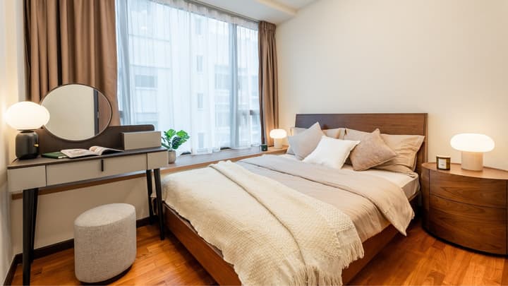 Bedroom with a large window, hardwood floors, a wooden queen bed with neutral bedding, and a modern gray dressing table with a round mirror.