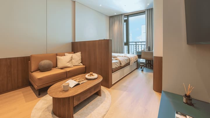 Wide view of a modern studio apartment with a queen bed and a separated tan leather sofa lounge area featuring a unique adjustable height coffee table, set near a large window.