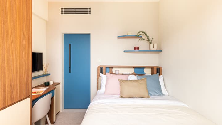 View of a compact studio featuring a queen-sized bed with wooden headboard, a pale blue door, a small desk/dressing area to the left, and floating blue accent shelves above the bed.