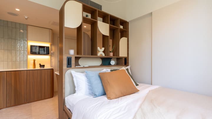 View of a queen bed with a wooden, open-shelf divider/headboard that separates the sleeping area from a modern kitchenette in the background.