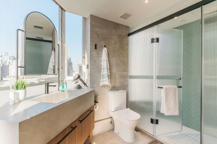 Modern bathroom featuring a concrete-style countertop and sink vanity, a toilet, and a walk-in shower with fluted glass doors and light blue-green herringbone tile, set against a large window with a cityscape view.