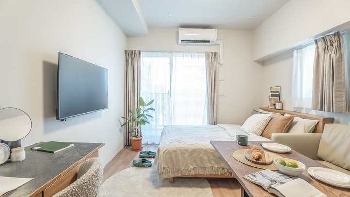 Wide view of a bright studio apartment with a double bed, a wooden table used for dining, a wall-mounted TV, and a work desk/vanity with a round mirror.