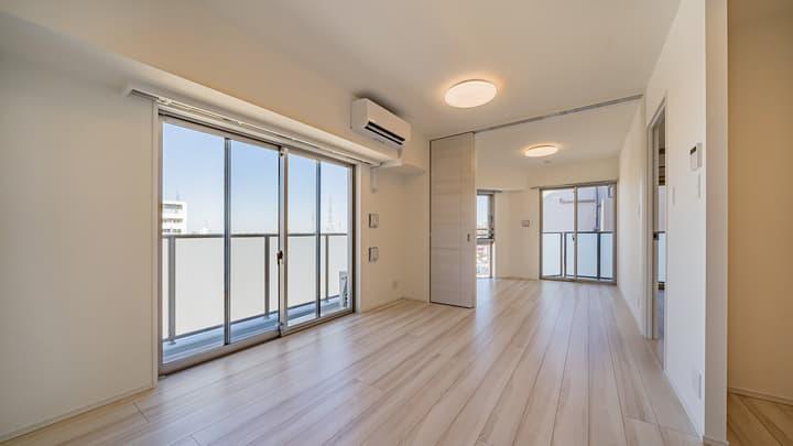 Long view of a spacious living and dining area with light wood-style flooring, a large sliding glass door to a balcony on the left, and a partial view of another room and balcony in the distance on the right.