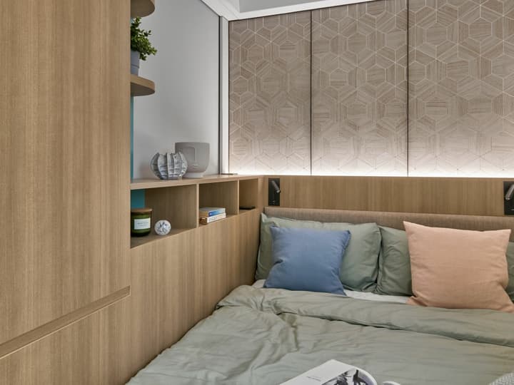 Close-up of a cozy double bed in a studio suite with pale green bedding and colorful pillows, featuring a wooden built-in headboard with integrated shelving, reading lights, and a geometric-patterned wall panel above.