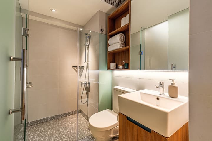 Modern bathroom in a premium suite featuring a square vessel sink on a wood-look vanity, a glass walk-in shower with gray terrazzo flooring, and wooden shelving above the toilet.