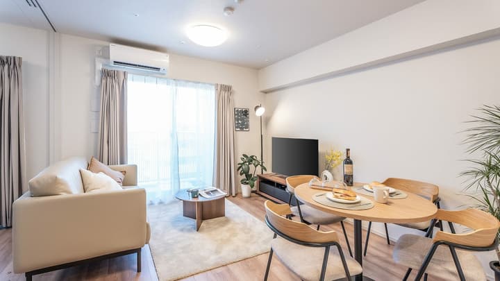 Minimalist living and dining room with light wood flooring, a beige leather sofa, a small round coffee table, and a dining set, next to a large sliding glass door leading to a balcony.