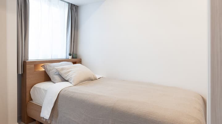 Compact bedroom with a single bed featuring a light wood frame, neutral bedding, and a large window with sheer and gray blackout curtains.