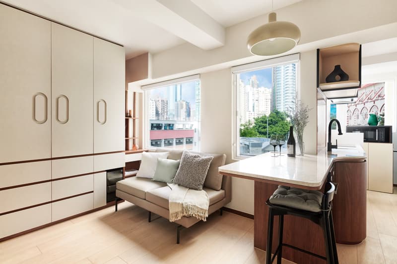 Contemporary, open-plan studio apartment featuring a sofa, sleek built-in floor-to-ceiling storage cabinets, a kitchenette with a breakfast bar, and large windows with a city view.