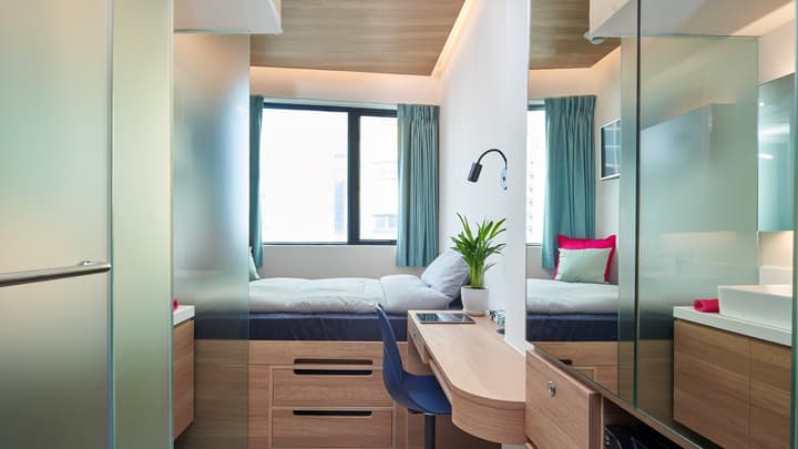 A compact studio apartment featuring a double bed with under-bed storage drawers, a light wooden desk, and large mirrors and frosted glass partitions that reflect the space and window.