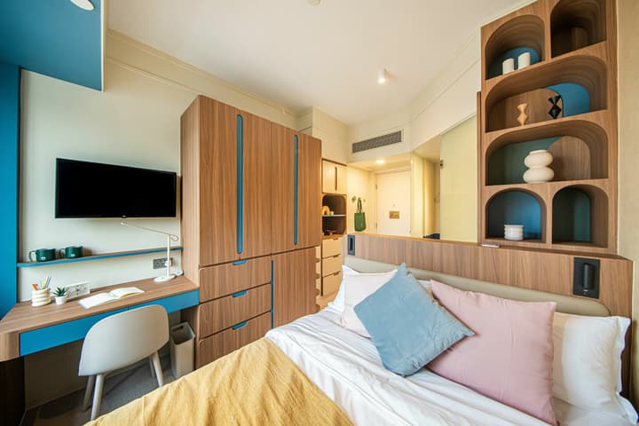 View of a modern, compact bedroom space with light walls and colorful accents. The room features a bed with pastel pillows, a built-in desk with a white chair and a wall-mounted TV, and floor-to-ceiling wood cabinetry with drawers for storage. A tall, decorative shelving unit with curved recesses stands to the right.