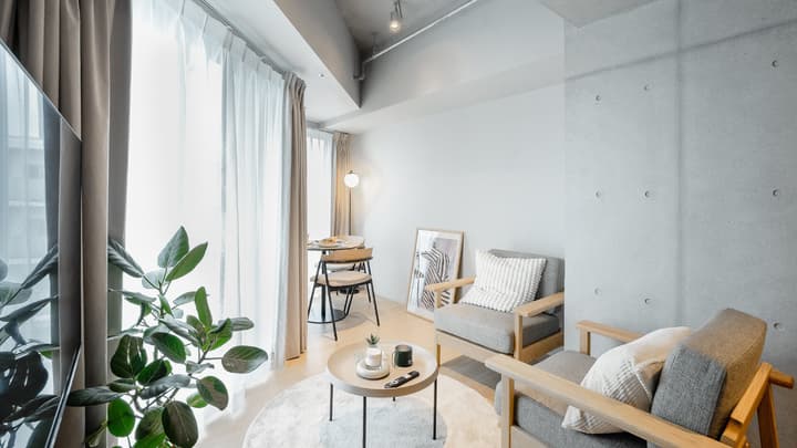Modern living space with two light wood and gray armchairs, a small round coffee table, a tall window with light curtains, and an exposed concrete accent column.