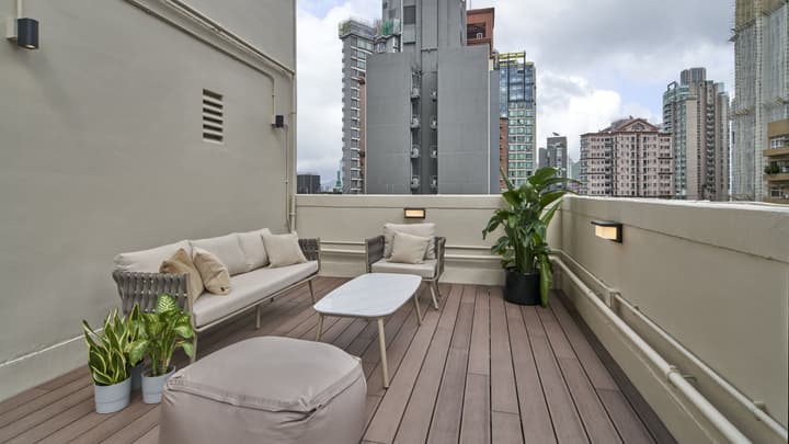 Modern rooftop deck with an outdoor lounge area including a sofa, armchair, coffee table, and potted plants, surrounded by tall city buildings.
