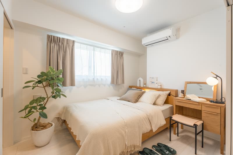 A fully furnished, bright bedroom with a wooden double bed, a matching wooden desk and mirror, a large potted plant, a window with beige curtains, and a wall-mounted air conditioning unit.