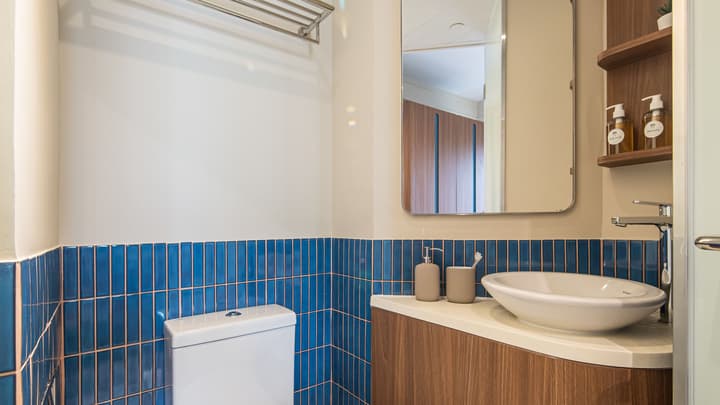 View of a compact, bright bathroom featuring a white toilet, a wall-mounted towel rack, and a sink vanity with a mirror above it, set against a wainscoting of blue mosaic tiles.