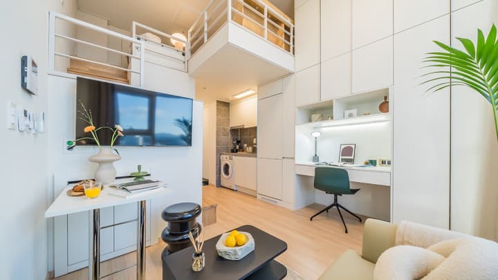 Wide view of a compact, bright loft studio apartment showing a living area, built-in desk, small kitchen, and an elevated sleeping area.
