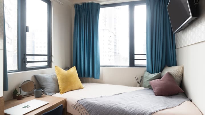 A compact student bedroom with a single bed, colorful decorative pillows, bright blue curtains on a large window, a small desk with a laptop, and a mounted television.