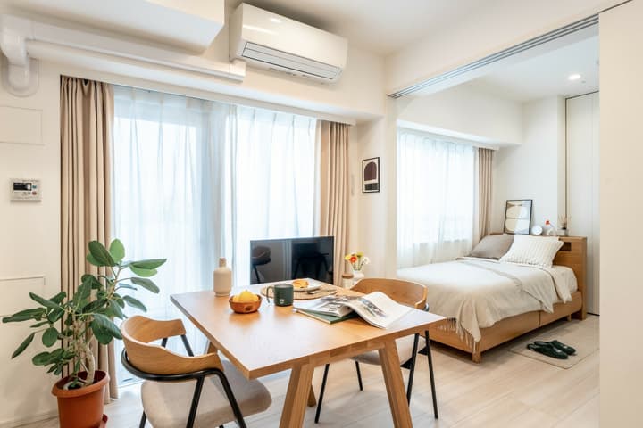 Open-plan living space with a wooden dining table and chairs, a television, and a visible bedroom area separated by a slight partition.