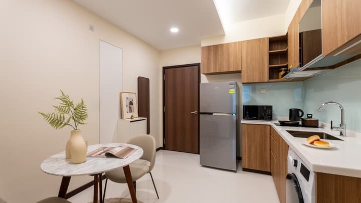 View of an apartment entrance area showing a dark wood door, a round marble dining table, and an adjacent kitchen with wood-look cabinets and a stainless steel refrigerator.