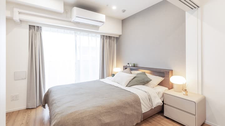 Furnished bedroom with a queen-sized bed, light grey textured accent wall, light wood-style flooring, nightstands with modern lamps, and large curtains covering a bright window.