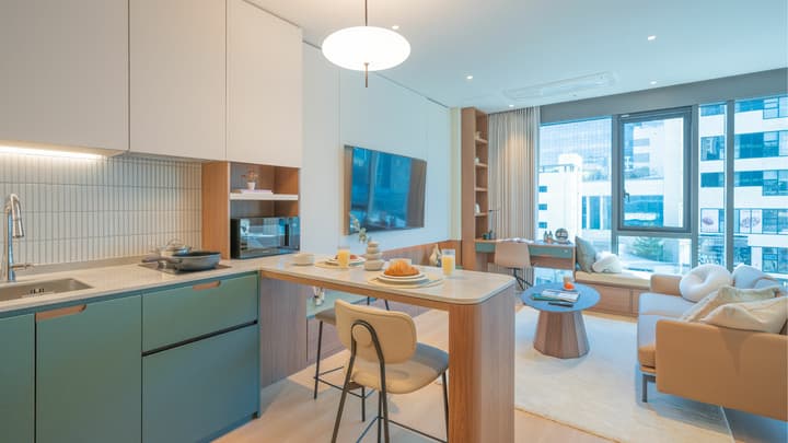 View from the kitchen toward the living area, showing the L-shaped green kitchenette, an extended counter bar used as a dining table, a wall-mounted TV, and a glimpse of the desk and window bench.