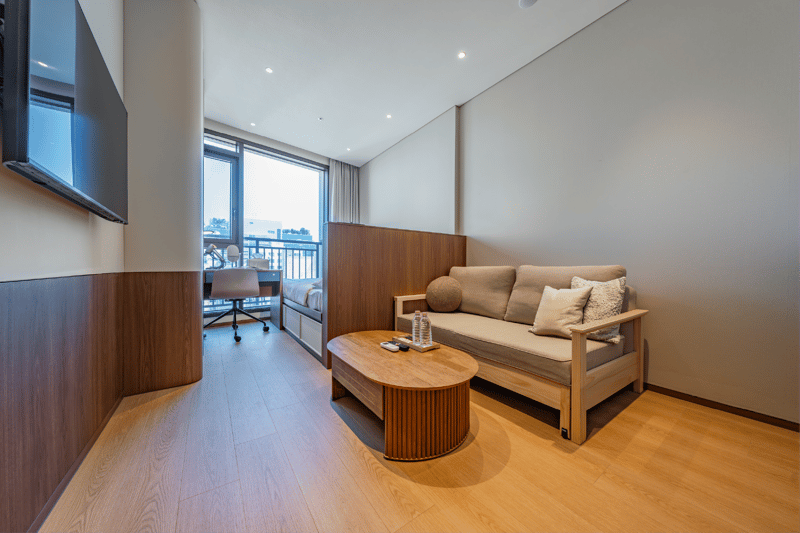 Wide view of a modern studio apartment with a queen bed and a separated sofa bed lounge area featuring a unique adjustable height coffee table, set near a large window.