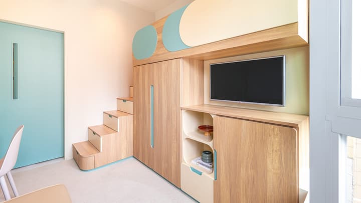 Interior view of a studio apartment showing a multi-functional wooden storage and bed unit with built-in stairs, a closet, and a mounted TV, next to a pale blue door.