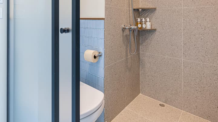 View into the shower area of a compact bathroom, showing large beige speckled terrazzo-style wall and floor tiles next to the toilet area behind a glass partition.