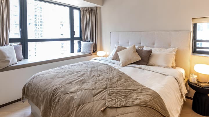 Bright master bedroom featuring a queen bed with a tufted headboard, a cushioned window seat, and a large window with city views.