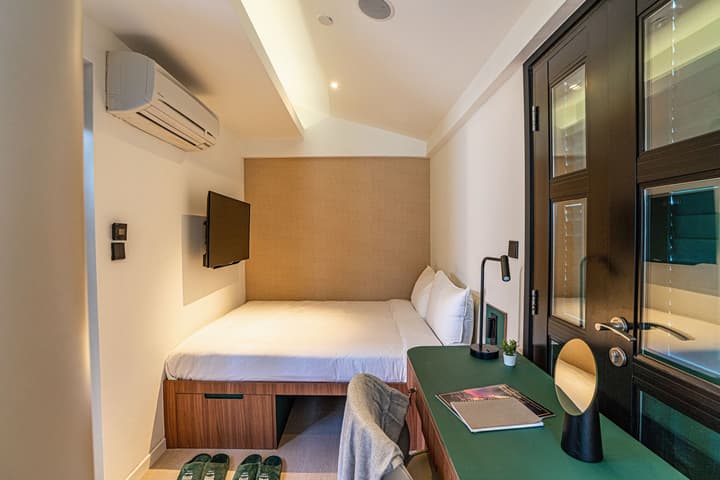 Sleeping area of a one-bedroom suite featuring a queen bed with woven headboard, a wall-mounted TV, and a built-in green desk with black accents.