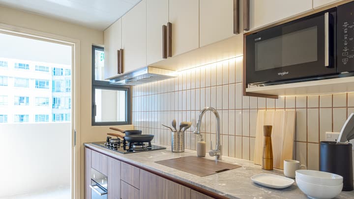 Modern kitchen with an induction cooktop, light wood and white cabinetry, a microwave, and vertical tiled backsplash, with a doorway leading to a balcony.